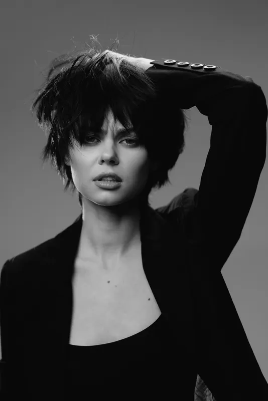 Black and white portrait of a young woman with short hair wearing a dark blazer, showing an intense and confident expression.