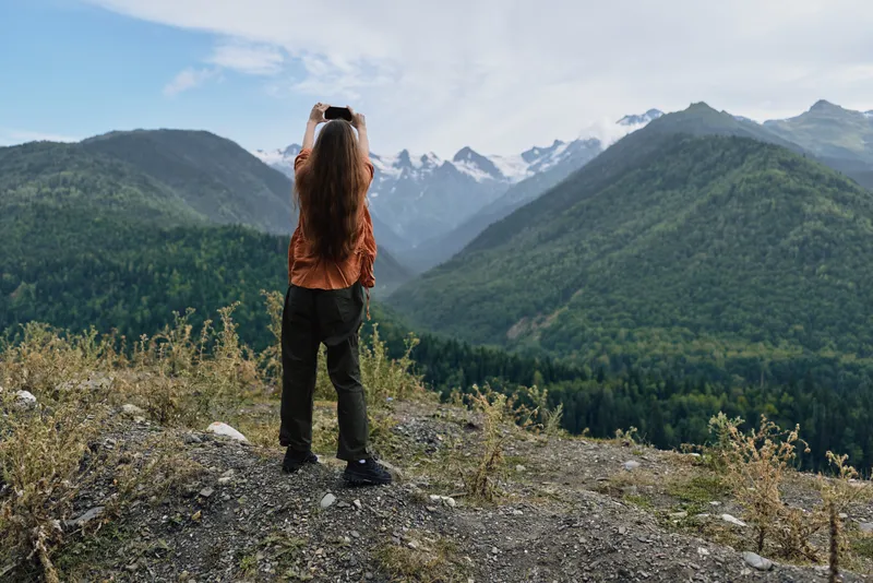 Person stands on a rocky overlook taking a photo of distant mountains, capturing a vast outdoor landscape and serene nature in a dramatic valley vista.