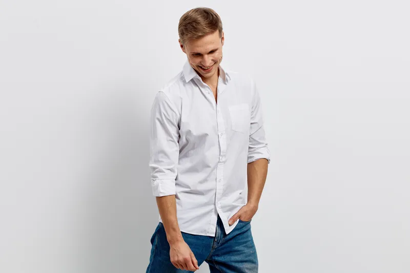Smiling young man with light brown hair wearing casual white shirt and blue jeans posing casually against plain white background studio portrait. People lifestyle concept