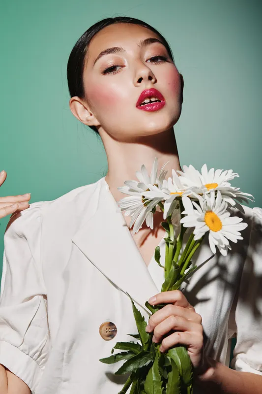 Fashion model with elegance holds white daisies close to her chest wearing a cream blouse on a muted green background highlights clean beauty with creative styling.