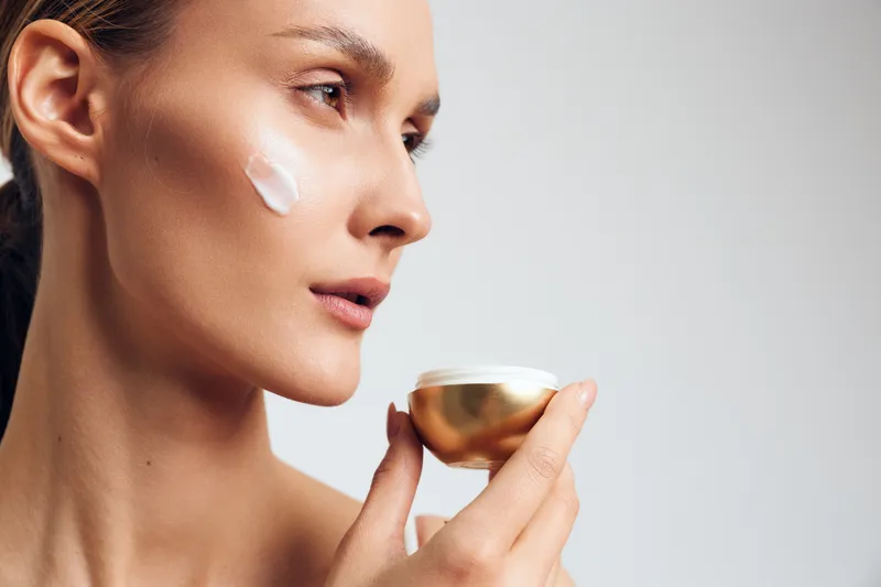 Young woman applies cream on face holds jar with skincare product has smooth skin natural beauty isolated on white background closeup profile portrait.
