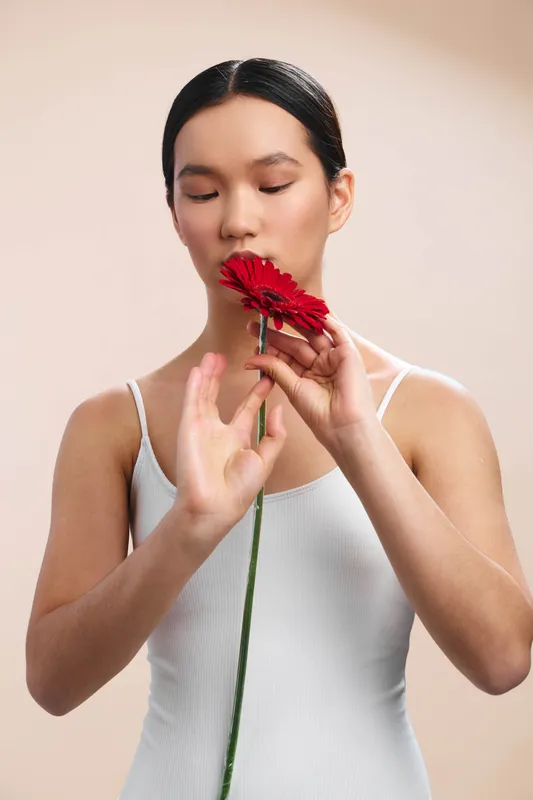 asian woman delicately holds a bright red flower near her lips, exuding calm focus and gentle grace in a soft studio setting, while light softly surrounds her and highlights serene beauty.