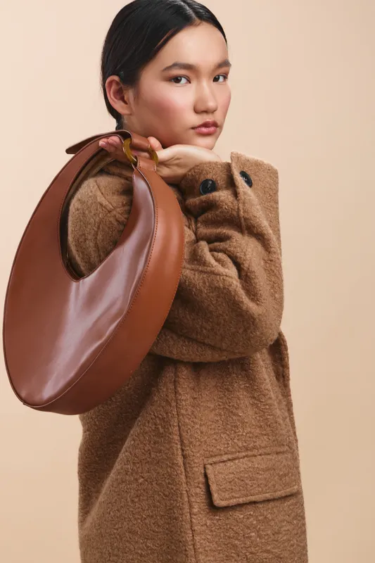 Asian woman exudes confident elegance in a chic winter coat, posing with a brown handbag. Soft studio lighting highlights texture and style, creating a refined fashion mood.