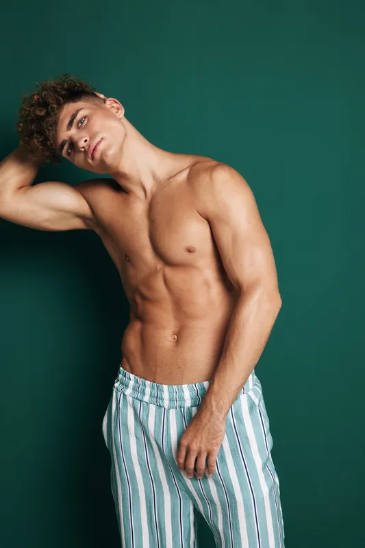 Young man with muscular body posing shirtless in striped pants against dark background. Fitness, toned physique and casual style with confident expression and curly hair.