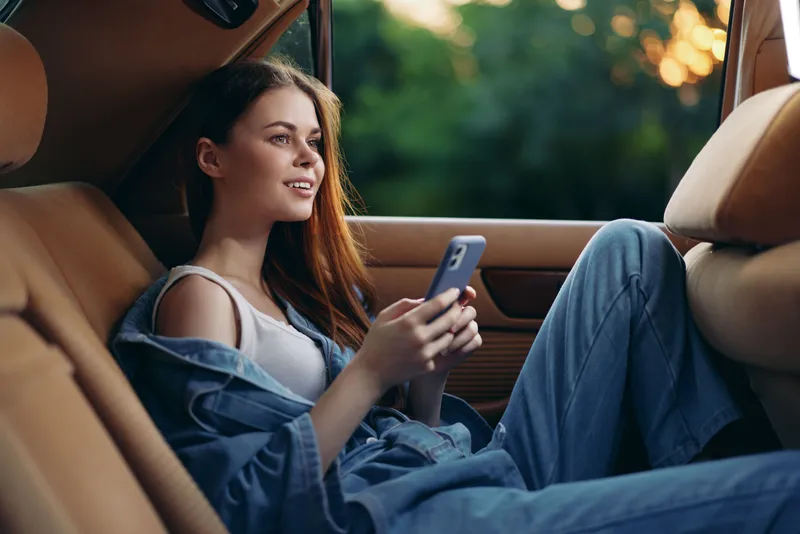 A young woman relaxes in the backseat of a car, wearing casual denim attire, smiling as she checks her phone. The lush green background adds a vibrant touch.