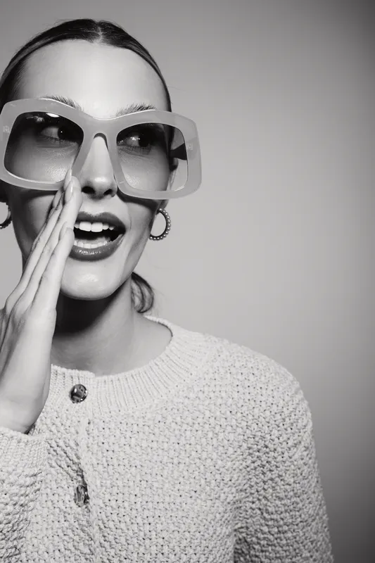 Young white woman with light skin and large sunglasses whispers with hand near mouth. Studio portrait in black and white with casual clothing.