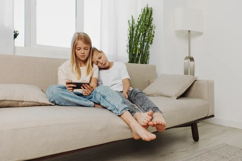 Two children sit barefoot on beige sofa playing on digital tablet in bright modern living room with plants and cozy cushions. Concept of family, childhood, and technology.