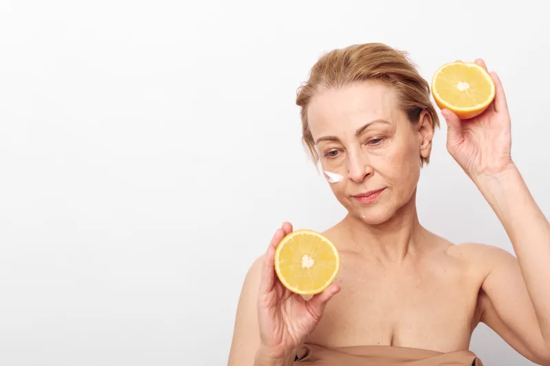 Mature woman with natural makeup holds two halves of fresh grapefruit near face, showing skincare cream on cheek. Concept of healthy skin, vitamin care, and natural beauty.