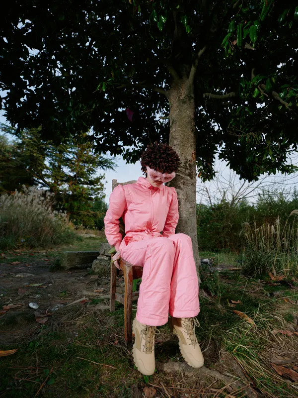 Fashion portrait of a person in a pink tailored suit seated under a tree in rugged outdoor setting, showcasing bold color, style details and confident feminine presence.