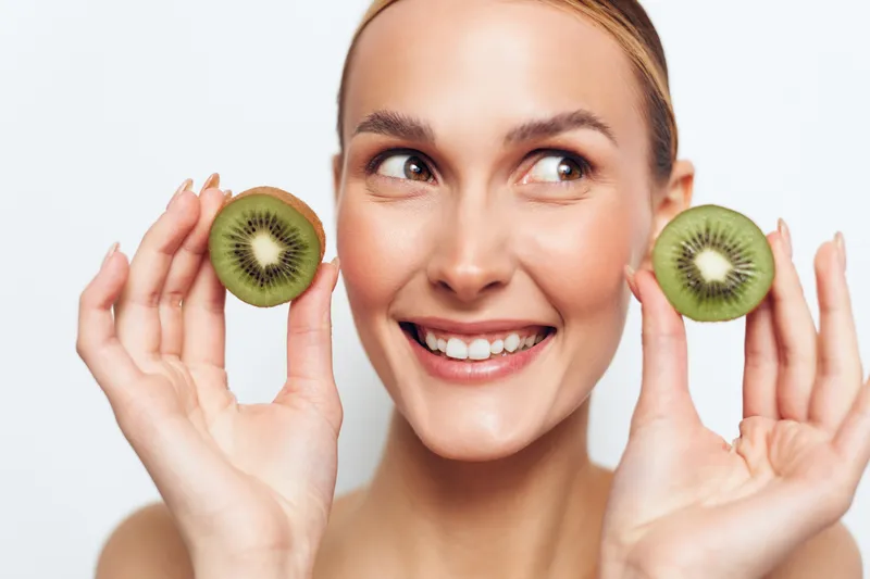 Woman holds kiwi slices near cheeks, smiling in fresh fruit skincare concept, healthy beauty portrait isolated on white background, natural light, copy space