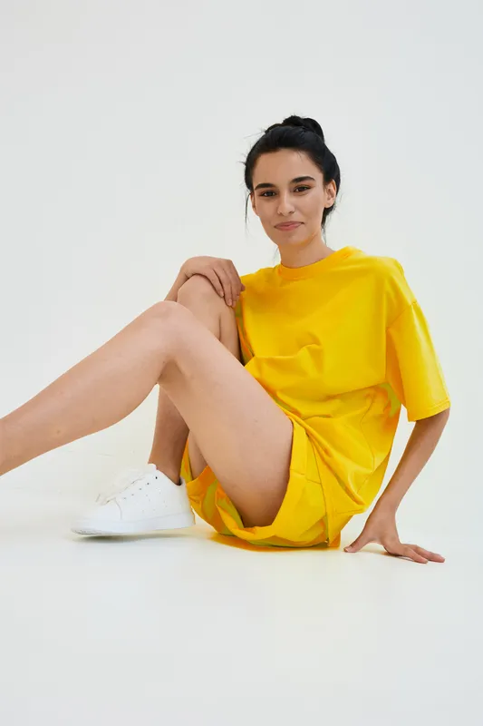 young woman in yellow casual outfit sitting on white floor, relaxed pose, minimalistic studio background, sporty style with white sneakers