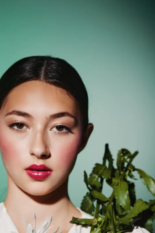 Close-up portrait of a young woman with dewy skin wearing vibrant red lipstick against a green background holding fresh leafy greens. Concept of beauty, fashion, and skincare creativity.
