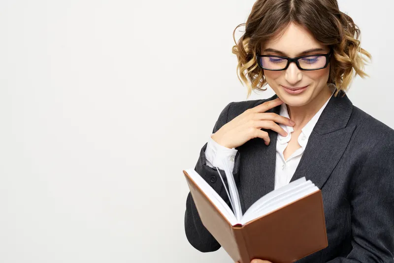 Женщина за работой с книгой в руке на светлом фоне, классический костюм, очки, голова