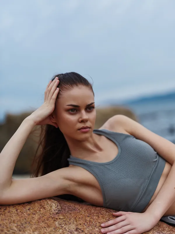Young athletic woman in a sports bra lounging on a rock by the ocean, showcasing confidence and strength in a serene, natural environment