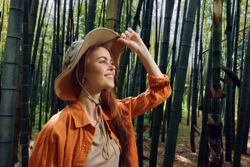Woman in a bamboo forest wearing a straw hat and orange jacket, smile and relaxed pose among tall green stalks, nature portrait capturing travel, joy and peaceful outdoor moment.