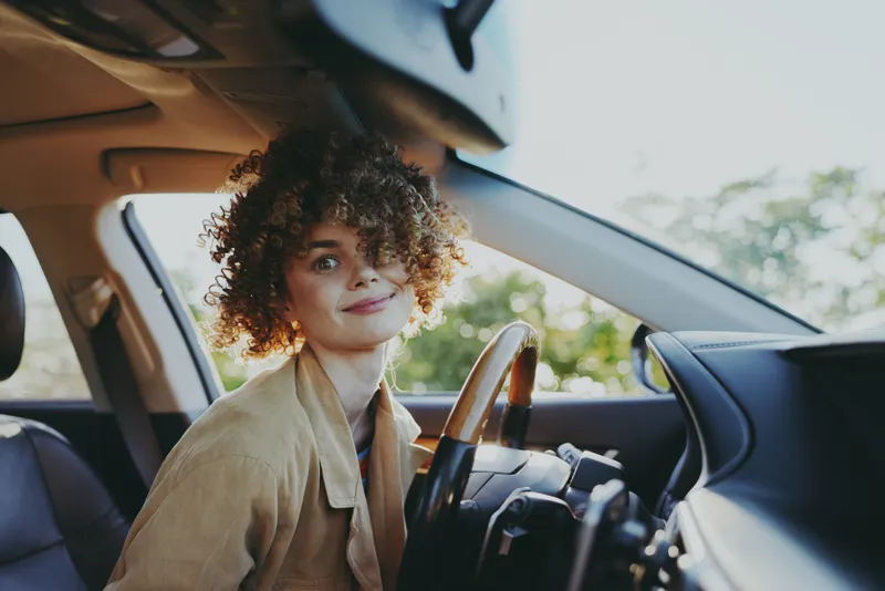 Young woman with curly hair smiles while sitting in a sunlit car, looking toward the camera, her beige jacket contrasting with dark leather seats and soft daylight highlighting the scene