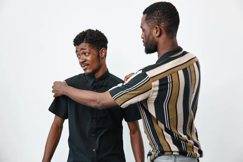 Two young black men with dark skin in casual clothes interacting on white background. One man comforts the other with hand on shoulder showing support and friendship.