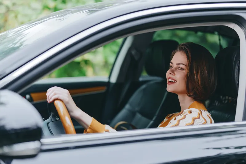 Portrait of a smiling woman driving a car, hands on the wheel, looking ahead in a bright outdoor setting.