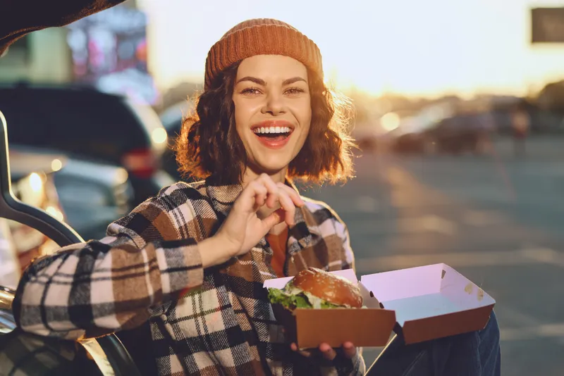 Woman with wide smile holding a takeout burger outdoors at golden hour, candid authenticity and joyful expression conveying mindful living and emotional storytelling in urban lifestyle.