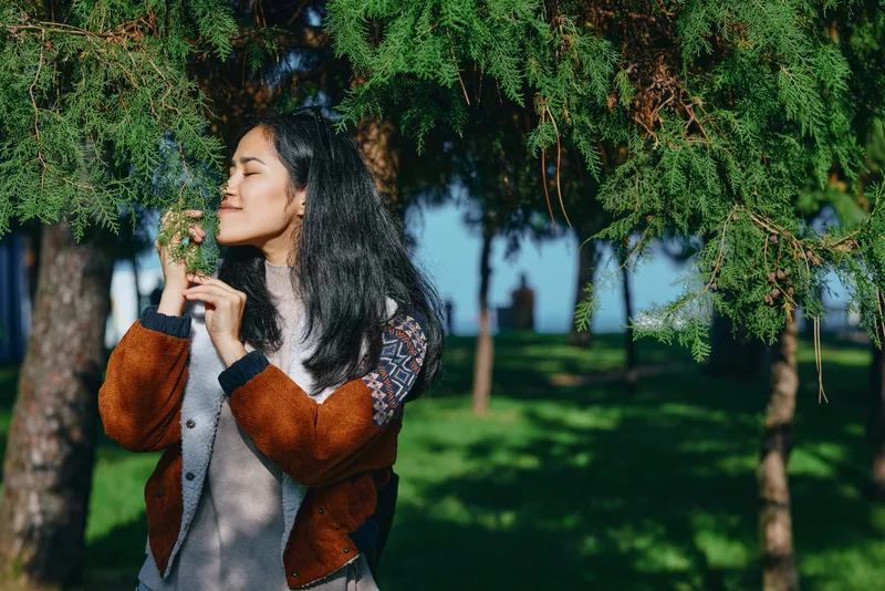 woman, tree, nature, outdoor, park, greenery, jacket, contemplative moment in a sunlit park as the woman touches a branch, gentle breeze, calm mood, candid scene among lush foliage and dappled light