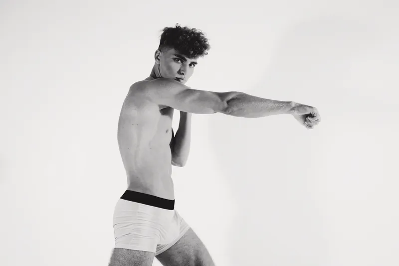 Young man in white shorts practicing boxing stance with focused expression on white background. Athletic male training, fitness and strength in studio workout.