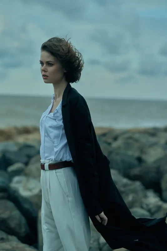 Windy ocean scene with a woman on the coast wearing a white dress and long black coat, showcasing moody fashion and sea breeze for editorial travel storytelling