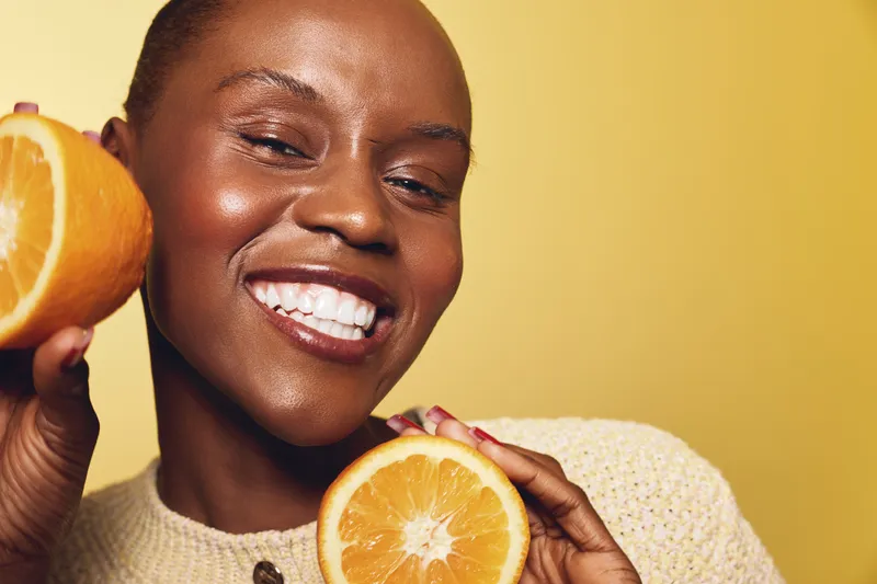 Happy smiling black woman with dark skin holding two orange halves in hands on yellow background. Concept of healthy lifestyle, vitamin C, natural beauty, and wellness.
