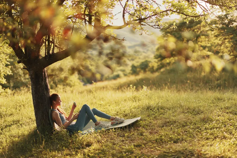 Young woman relaxing under a tree in a serene natural setting, enjoying a book in the warm sunlight, evoking feelings of peace and tranquility amidst nature