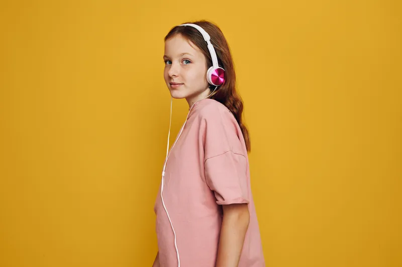 Teenage girl with red hair wears pink headphones and a loose pink t-shirt standing against a bright yellow background. Side view portrait in studio lighting.
