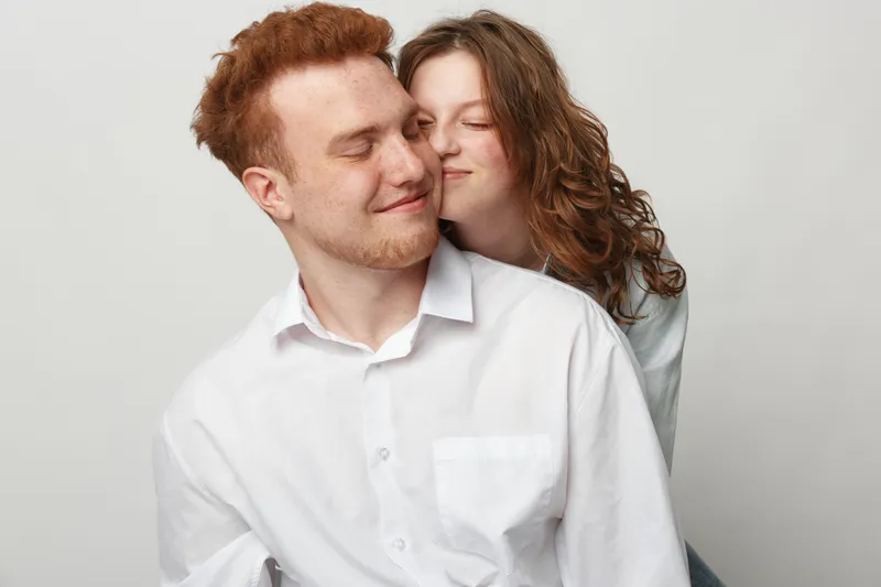 Happy young couple with red hair and curly hair embraces gently, smiling with closed eyes. Casual clothing and light background create warm, intimate, and loving relationship concept.