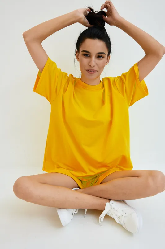 Young woman with dark hair tying a bun, wearing bright yellow t-shirt and white sneakers, sitting cross-legged on a white seamless background, casual and relaxed pose.