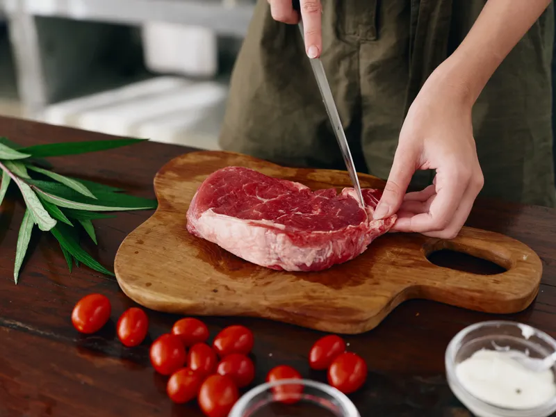 Женщина с ножом в руке нарезает свежее стейковое мясо для жарки на кухне; на столе соль, перец и другие специи, красные помидоры черри и зелень, приготовление ужина
