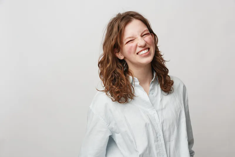 Young woman with curly hair wearing white shirt smiles with eyes closed against plain background. Concept of happiness, joy, casual style, and natural beauty.