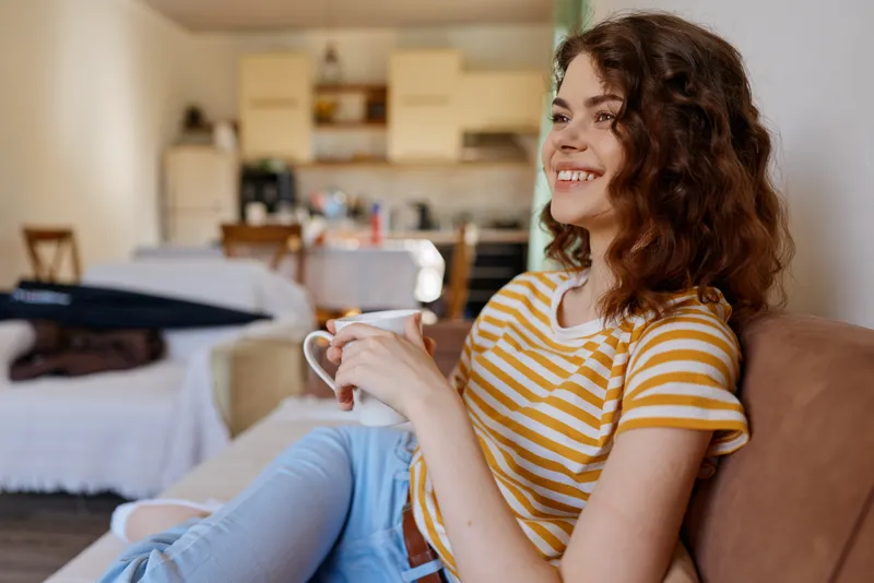 Woman relaxes at home, smiling, holding a mug in a striped shirt, casual lifestyle scene in a cozy living room with open kitchen in the background, natural light, comfortable mood and bright ambiance