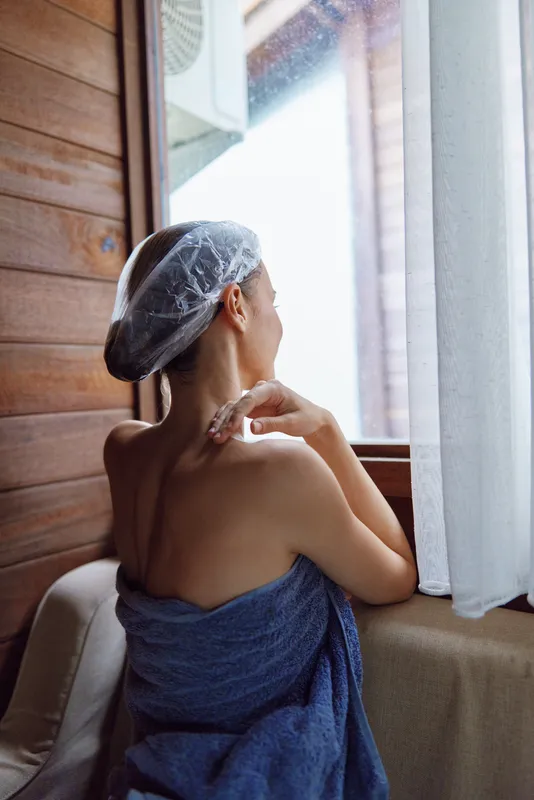 Woman wrapped in towel with hair cap resting by window in cozy wooden interior, natural light illuminating peaceful morning moment with calm atmosphere and warm tones.