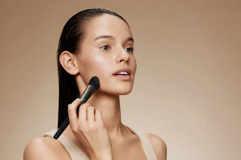 A young model applying makeup with a brush, showcasing natural beauty and confidence, against a soft beige background, perfect for beauty and skincare projects