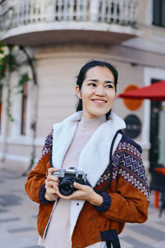 Smiling woman outdoors wearing a cozy jacket and fleece collar holds a camera ready to shoot, urban street scene, candid lifestyle photography moment
