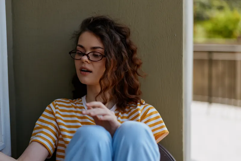 A contemplative woman with curly hair and glasses sits against a wall in a sunlit home, wearing a striped tee and blue jeans, conveying introspection, calm, and daily life.