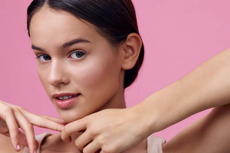 Young model with a natural look posing against a soft pink background, showcasing minimal makeup and a confident expression