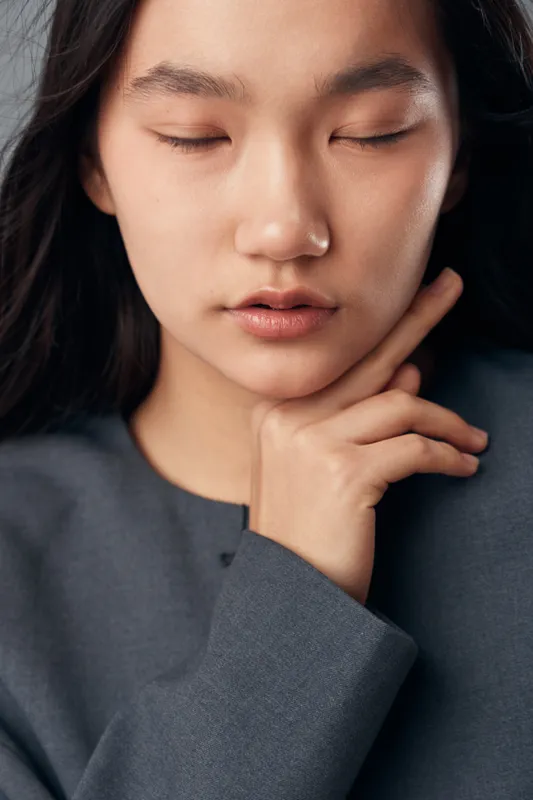 Young woman with closed eyes touching face gently, calm expression and natural makeup. Portrait of peaceful female model with dark hair and soft skin in casual grey clothing.