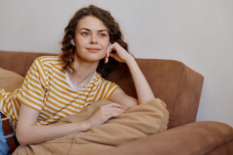 A young woman lounges on a sofa in a striped yellow t shirt, relaxed with a soft blanket nearby, conveying casual comfort and quiet contemplation.