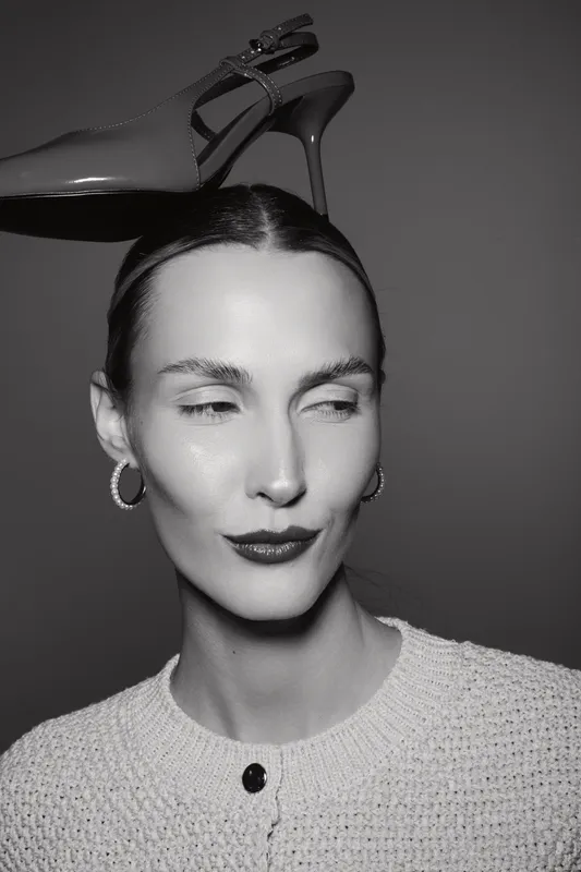 Caucasian woman with light skin balances high heel shoe on head, wearing hoop earrings and knitted sweater. Concept of fashion, style, and playful elegance in studio.