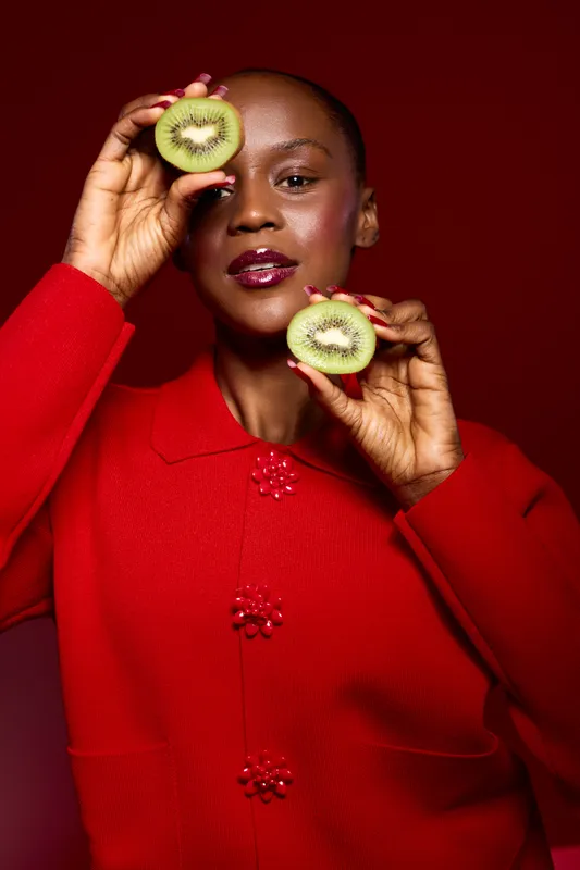 Portrait of a confident black woman with dark skin wearing a red sweater holding two halves of kiwi fruit near her face against a red background. Concept of healthy lifestyle and natural beauty.