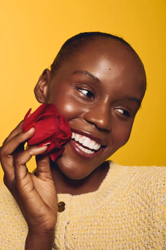 Smiling dark skinned African woman holding red rose near face looking sideways. Close-up portrait on yellow background. Concept of beauty, happiness, natural skincare, and floral freshness.