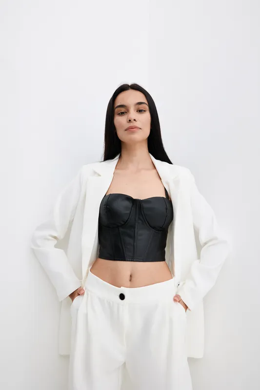 confident woman wearing white suit and black corset stands with hands in pockets against white background, showcasing modern fashion style and minimalistic look.