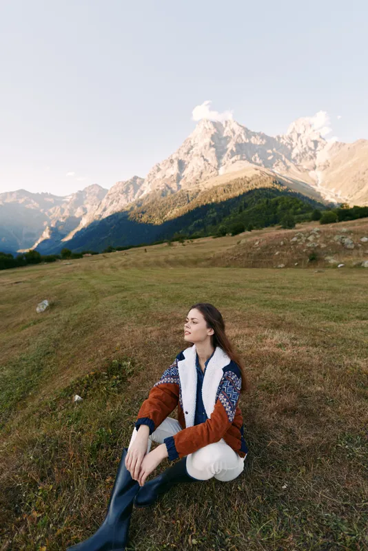 Woman sitting in meadow with mountains behind, portrait of travel and nature scene in autumn, cozy sweater and peaceful landscape for outdoor leisure and scenic horizon.
