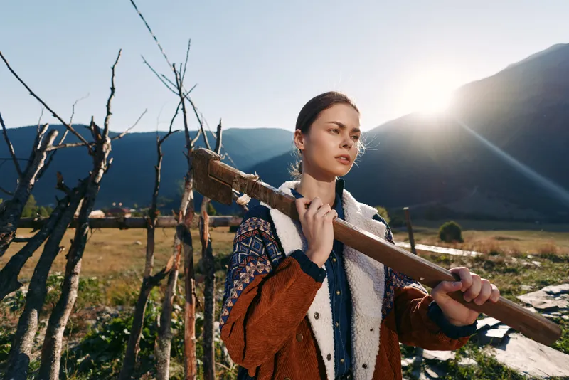 Woman with axe over her shoulder in a rural field near mountains and bright sun, wearing patterned jacket and fleece, portrait of farm work, outdoor lifestyle and resilience.