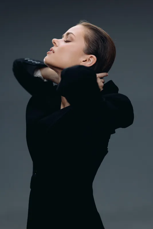 Elegant woman in black dress posing with closed eyes and serene expression on gray background.