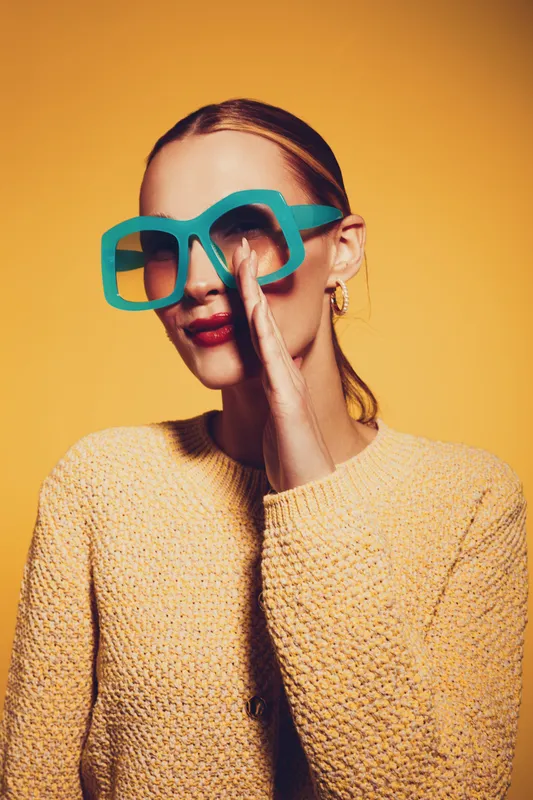 Young woman in oversized turquoise sunglasses and yellow sweater poses against yellow background. She smiles subtly with hand near mouth. Concept of fashion, style, and colorful portrait.