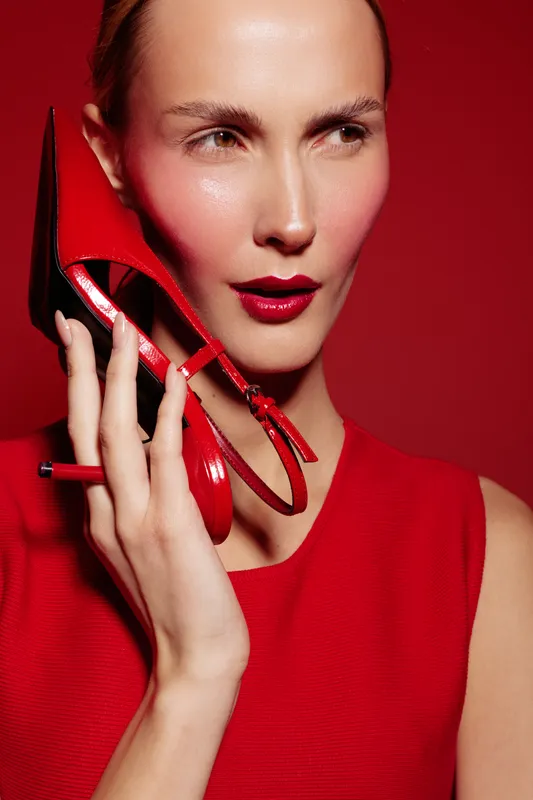 Stylish woman in red dress holds red high heel shoe near face on red background. Concept of fashion, beauty, glamour, and elegance with bold makeup and confident expression.